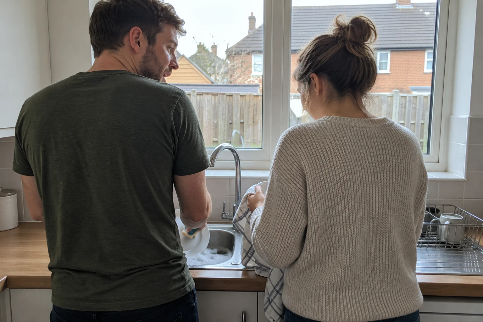 A couple standing side by side at a kitchen sink, washing up and talking